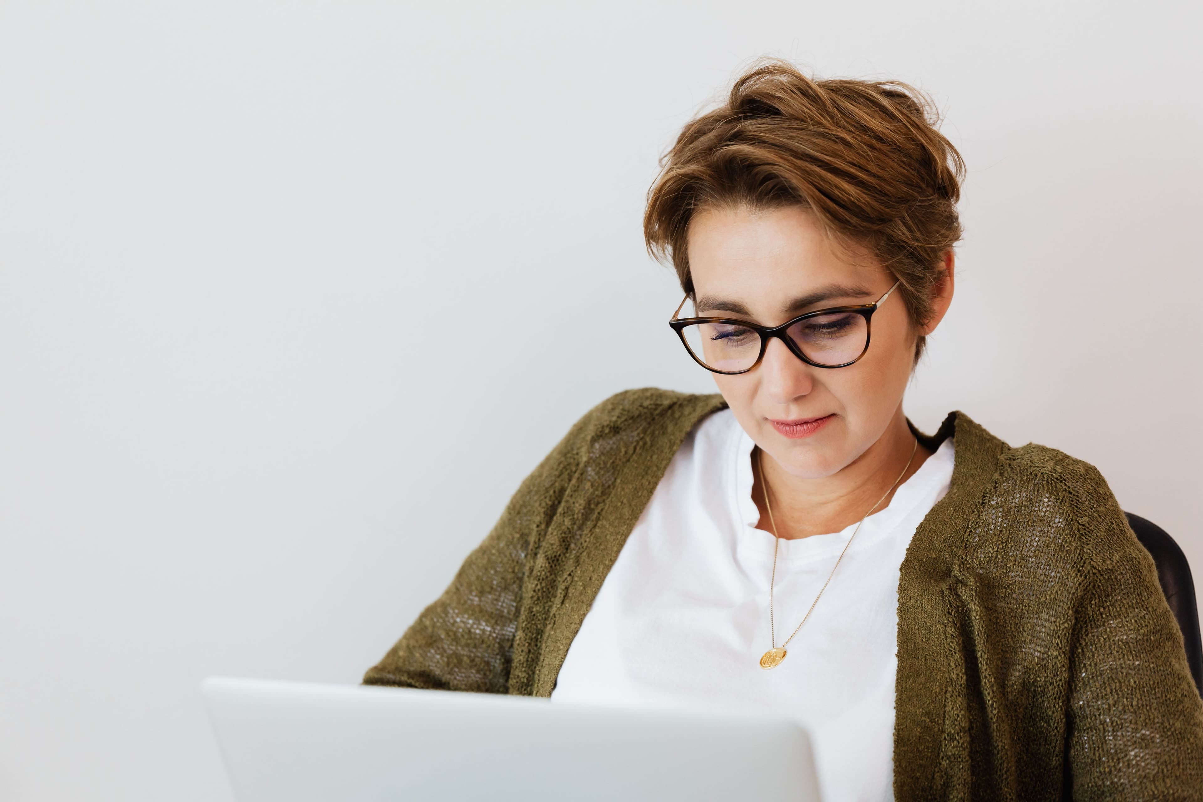 Smiling adult short haired female in stylish clothes and eyeglasses using laptop in light office space